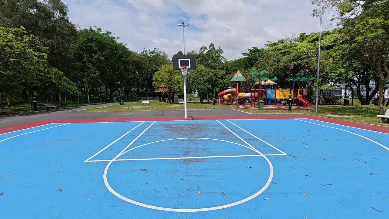 An empty basketball court in a tropical park, surrounded by lush green trees under a bright, sunny sky. Perfect weather.