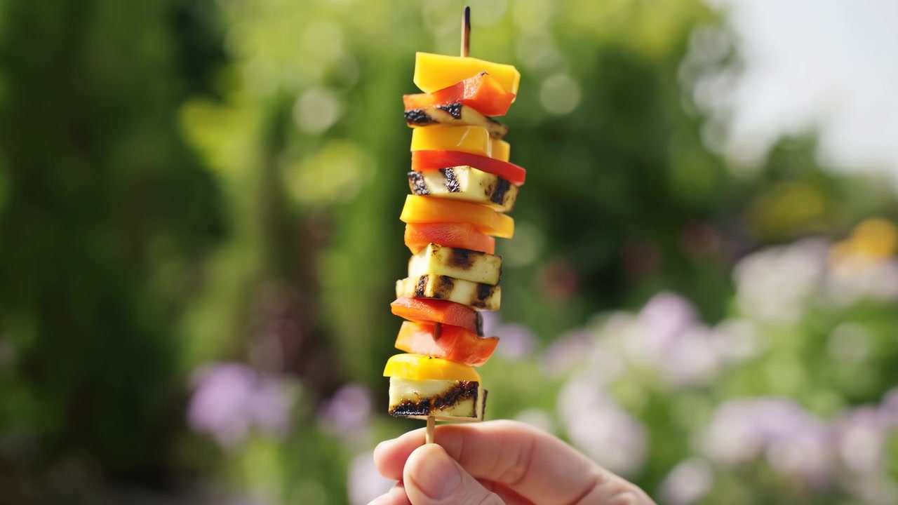 Primo piano di una mano che tiene uno spiedino verticale di pezzi di frutta mista in primo piano, con uno sfondo di giardino leggermente sfocato, vegetazione, fiori ed elementi naturali.
