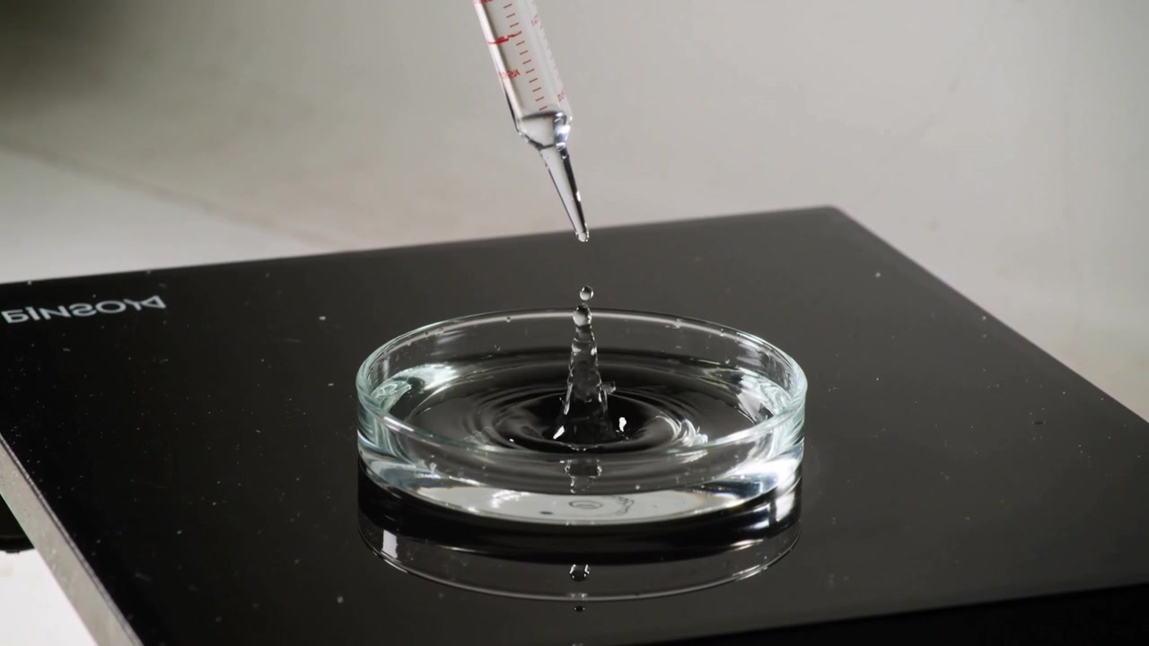 Una pipetta graduata rilascia gocce in una capsula di vetro su una superficie nera, formando una gocciolina e studiando le increspature.