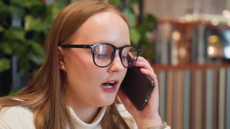 close up joyful young woman with glasses holding phone to ear enjoying lively chat with friend while sipping coffee seated indoors with warm ambiance and greenery in background