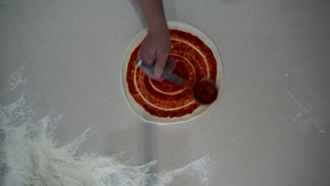 top down view of a chef making pizza on countertop with flour. overhead view of professional cook putting ketchup and salami on pizza dough on the table