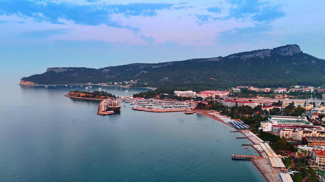 Flying along the beautiful beachline with greenery in Kemer, Turkey. Approaching the yacht club with multiple boats. Green rocks at backdrop