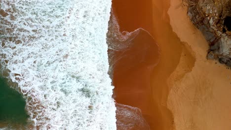 Aerial-shot-of-a-wave's-frothy-crest-breaking-along-the-shoreline,-The-sea's-lace-like-foam-meeting-the-beach