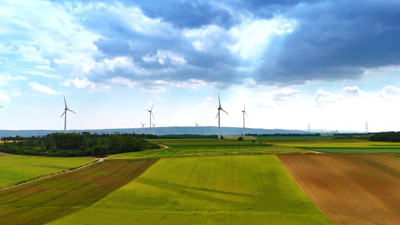 Flight over the mowed green field approaching the wind turbines. Grey thick cloudscape in the sky over the wind farms
