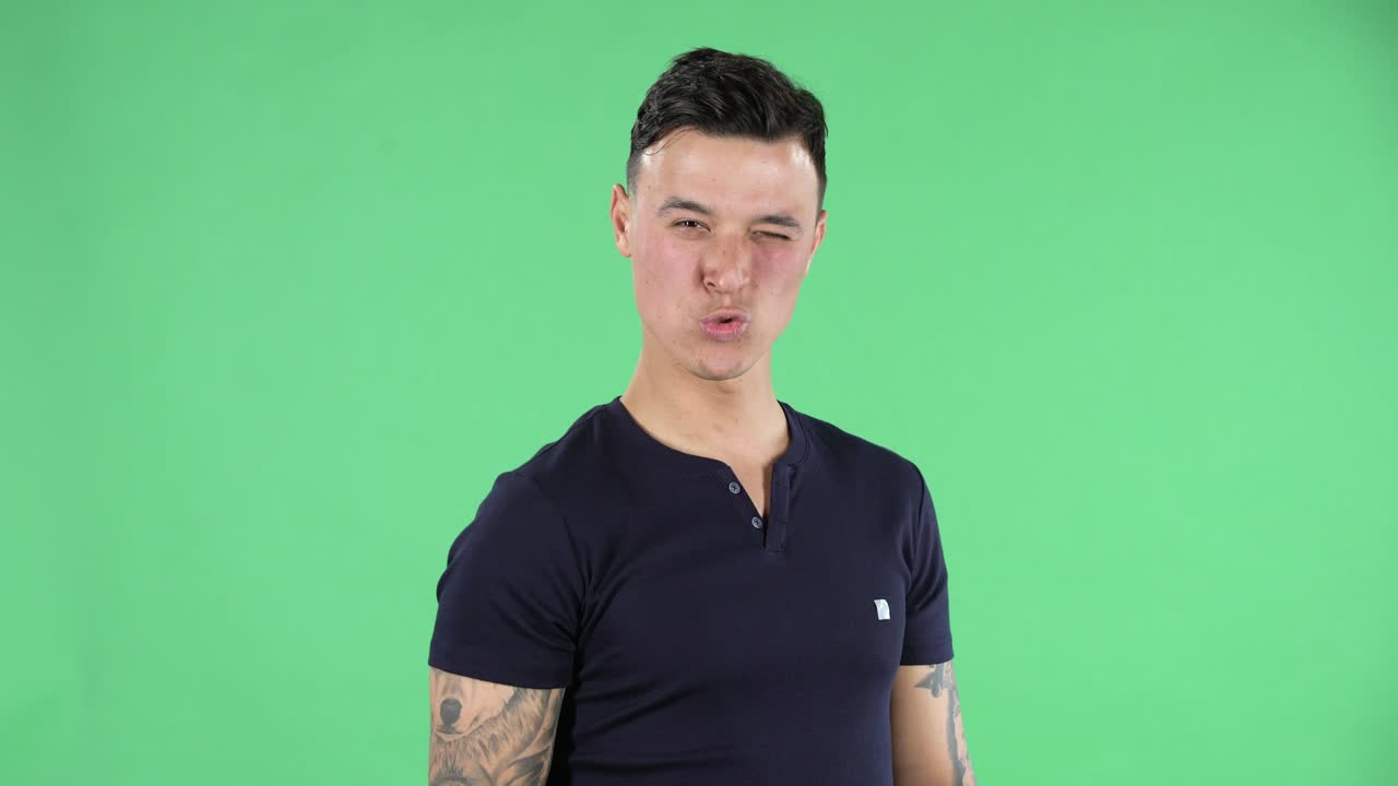 Portrait of a handsome young guy looking straight smiling and showing a Korean heart gesture, kissing and winking. Green screen