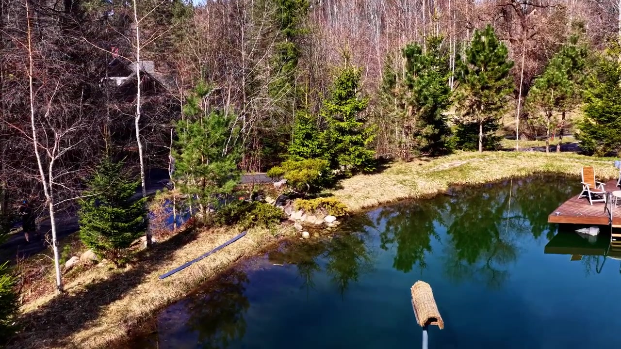 Drönarbild över en liten sjö med brygga och en central fågelholk i ett fridfullt landskap.