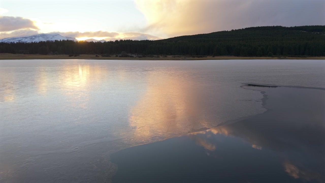 Arjantin, Chubut, Esquel yakınlarındaki Laguna La Zeta'nın donmuş yüzeyi, Patagonya ormanlık alanıyla çevrili kış buzunun üzerinde gün batımı renklerini ve dramatik bulutları yansıtıyor, drone geri çekilme çekimi
