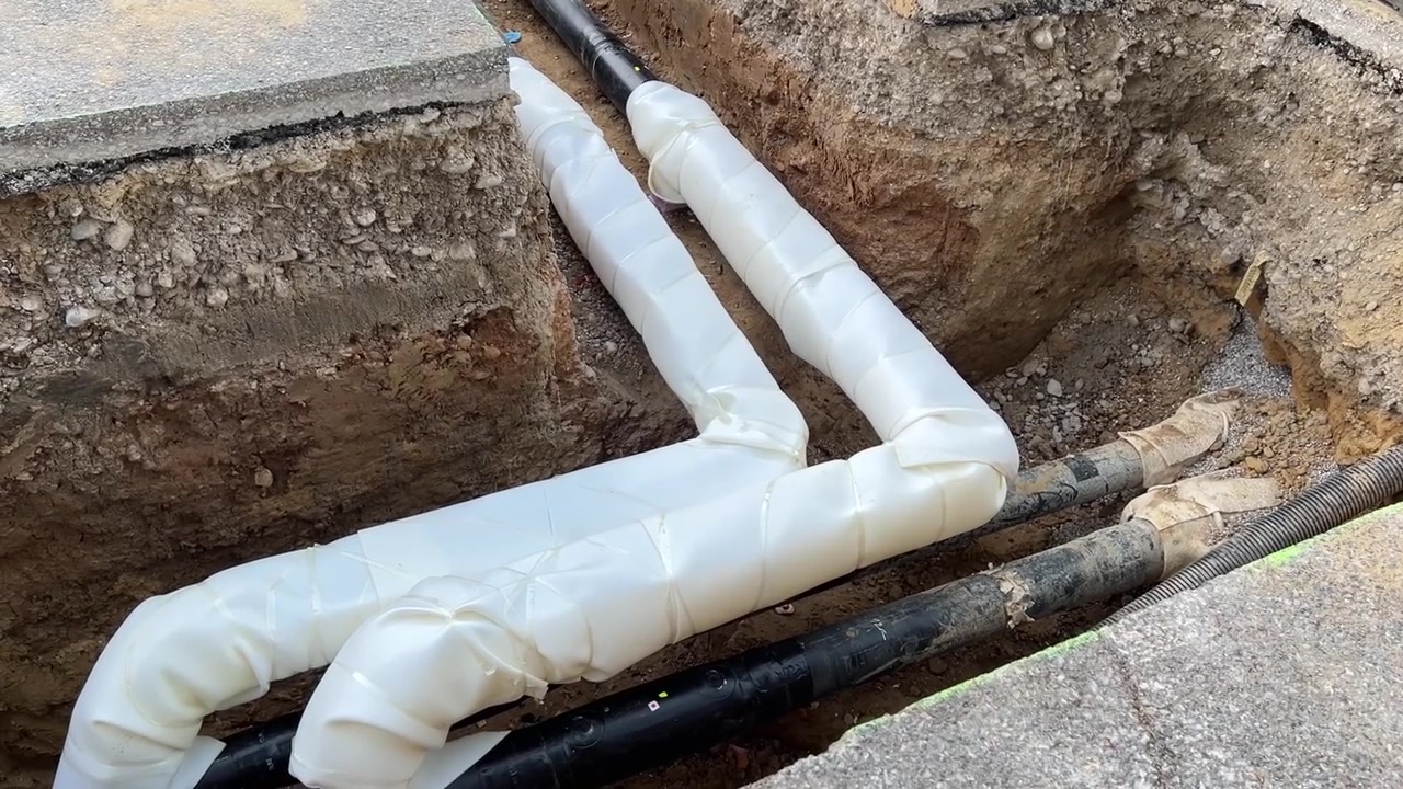 Open trench showing a 90-degree tee branch in pre-insulated district heating pipes, the junction wrapped in additional white insulation. Energy infrastructure and heat network installation, Germany.