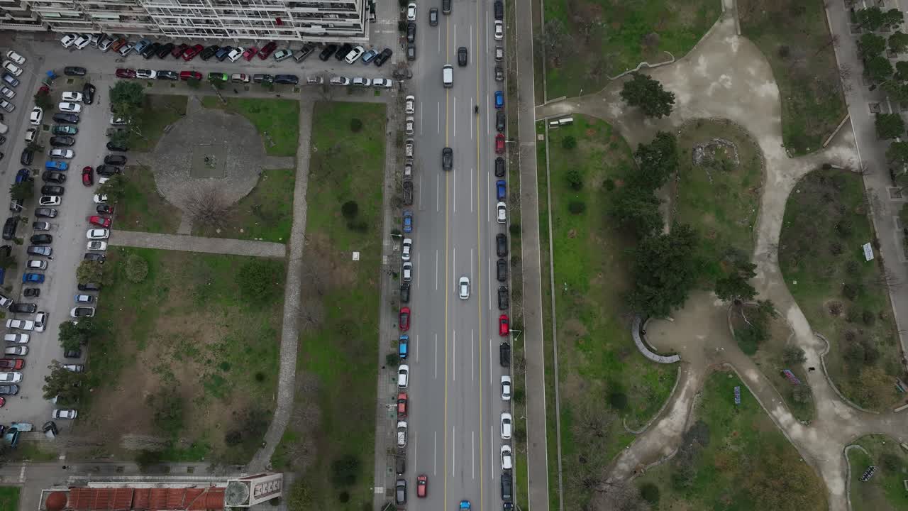 Drone view of Thessaloniki downtown, Greece, busy city streets, moving traffic, parked cars, urban buildings and green park spaces from top-down aerial view.