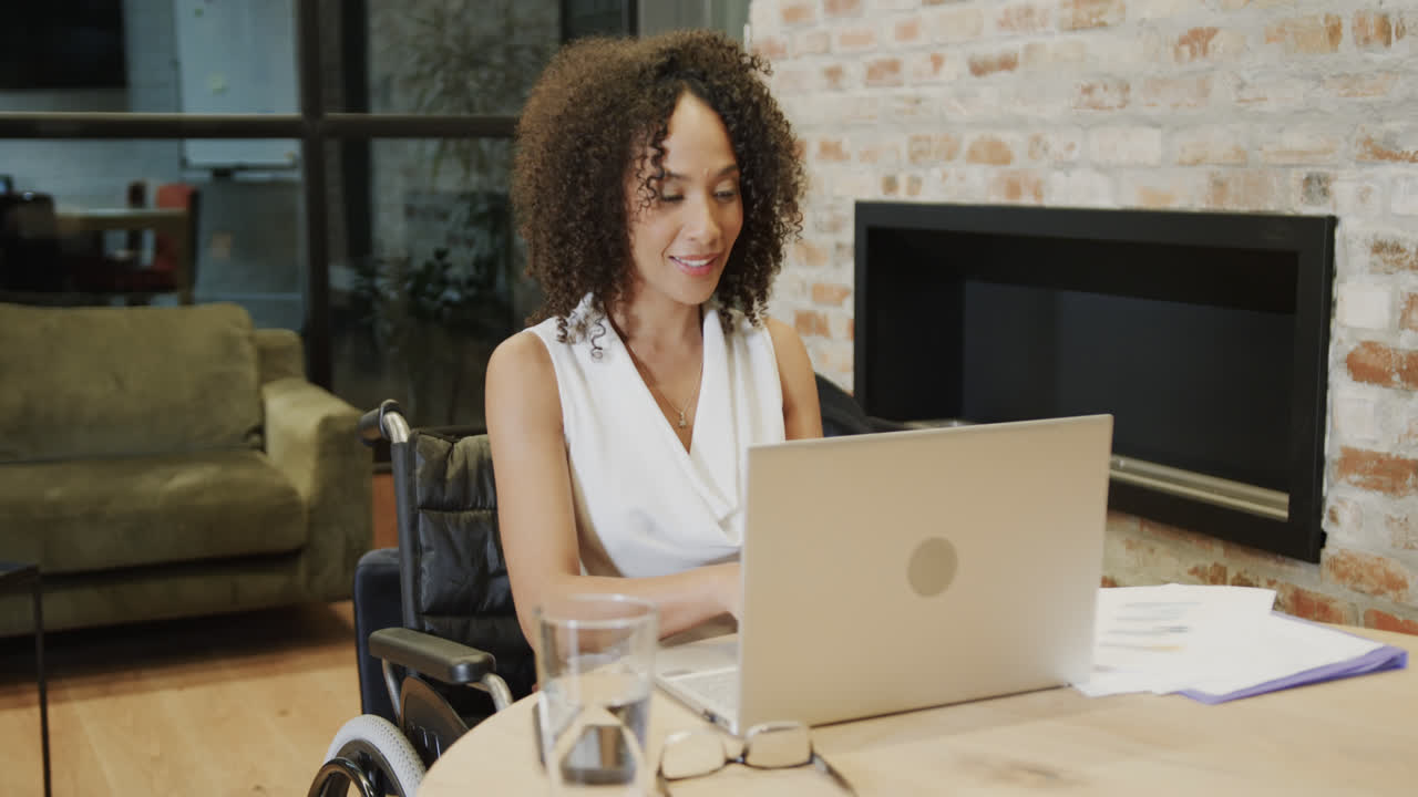 Felice donna d'affari birazziale in sedia a rotelle che usa un laptop alla scrivania in ufficio, al rallentatore.