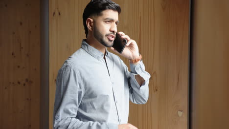 talking on smartphone, indian man standing by window in business casual attire