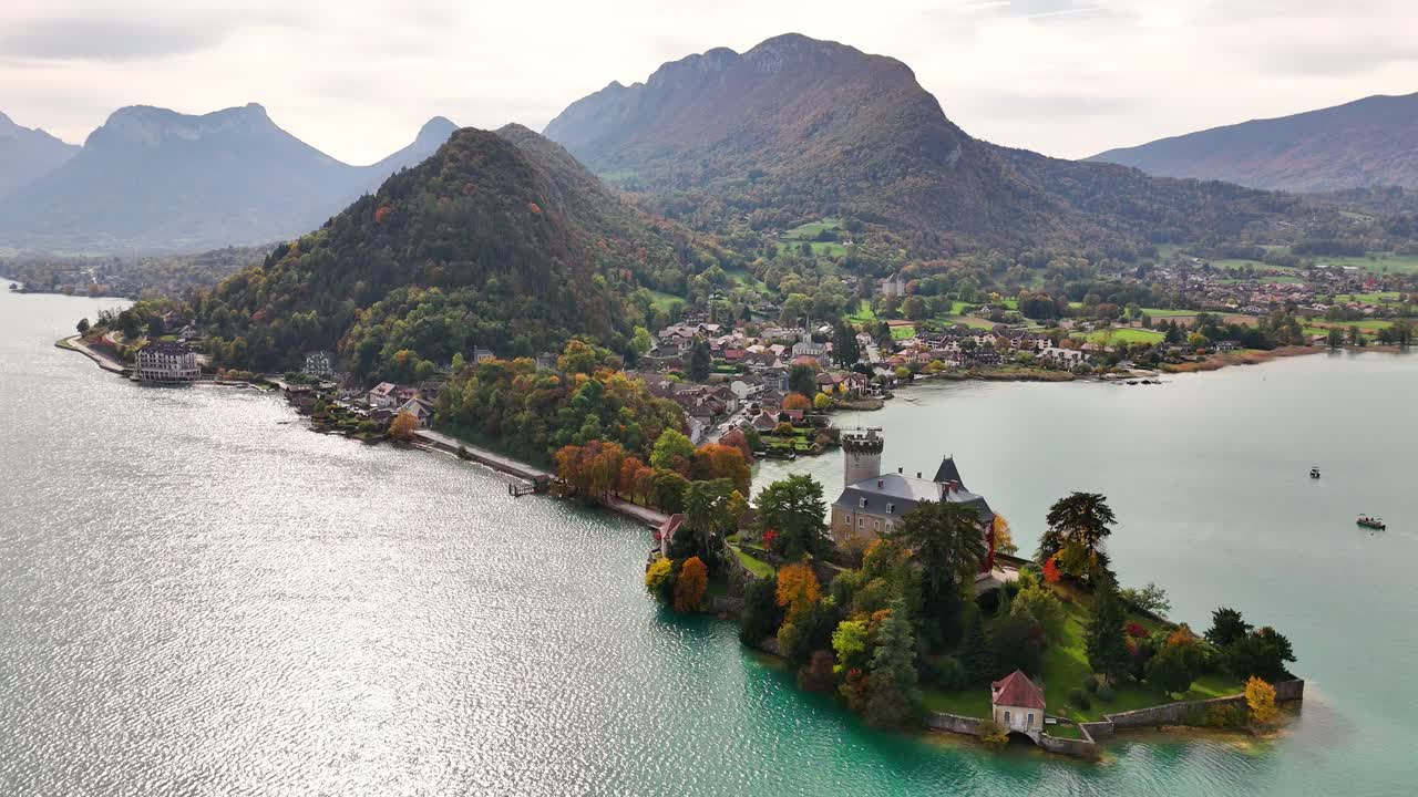 Aerial view of Chateau de Duingt in France surrounded by a calm lake, colorful autumn trees, a narrow peninsula, and nearby village with dramatic mountains forming a striking backdrop