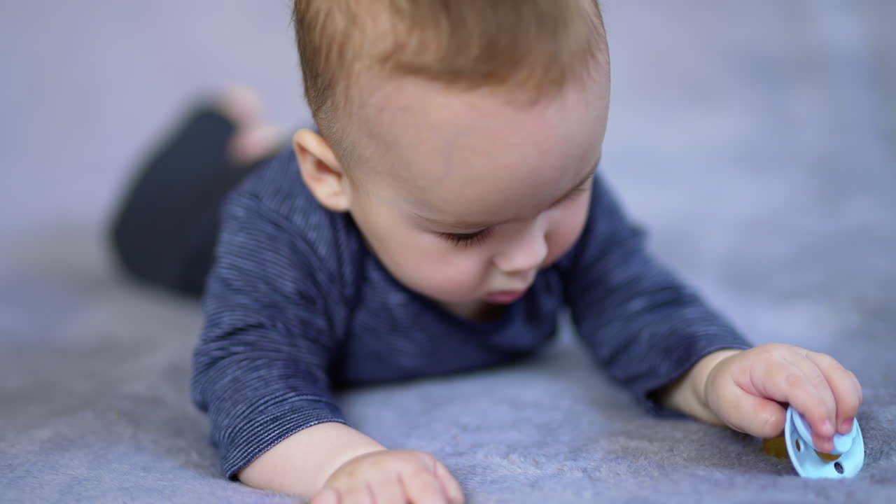 Adorable little baby boy lies on belly and takes a pacifier lying beside him. Sweet kid looks up and starts to smile cutely. Close up. Blurred backdrop.
