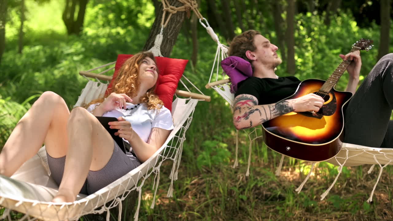 Couple lying on hammocks, talking and laughing. Young man playing guitar, woman smiling, working with tablet and stylus. Greenery around. Glamping. Slow motion