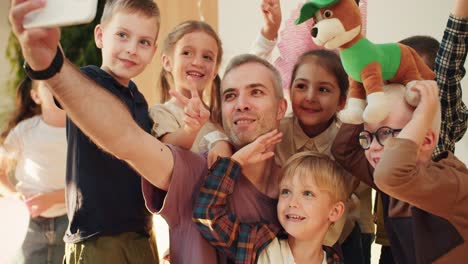 un grupo de niños preescolares felices junto con su maestro, un hombre de cabello gris en una camiseta púrpura, toman una selfie en su lección en un club para preparar a los niños para la escuela