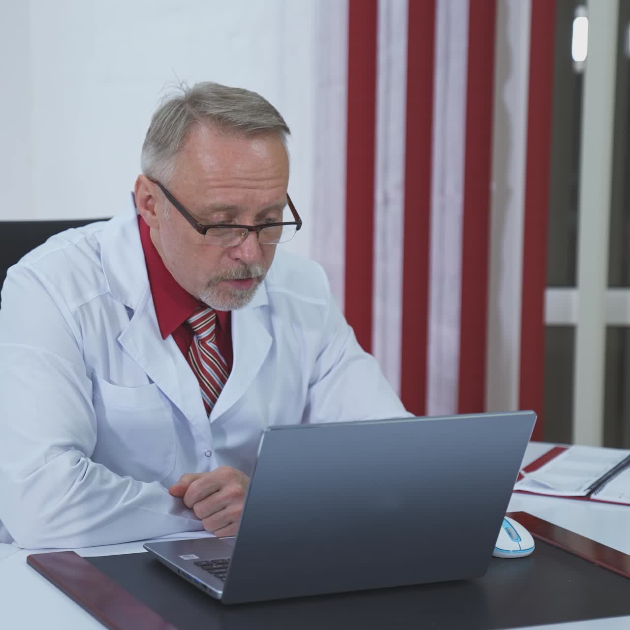 Professional doctor talks to distant patient. Male physician in white medical uniform speaking through video chat while looking at laptop in clinic