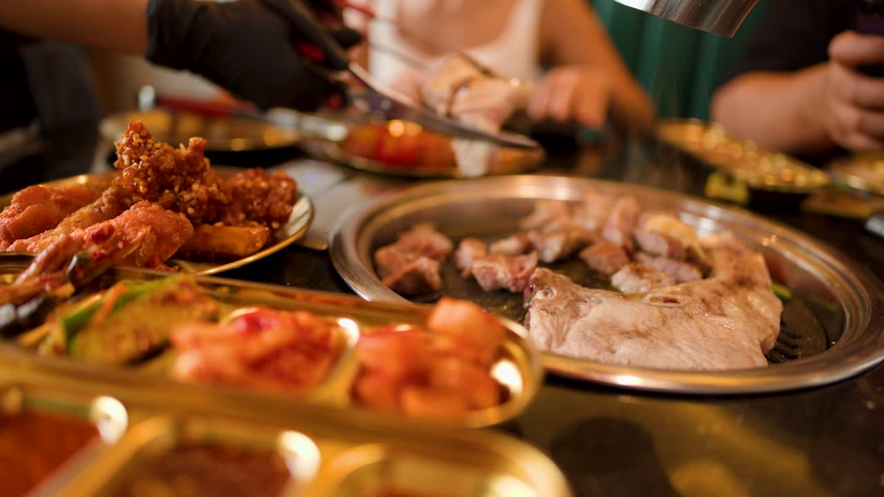Gloved hand cuts meat with scissors over grill, surrounded by side dishes, warm indoor lighting