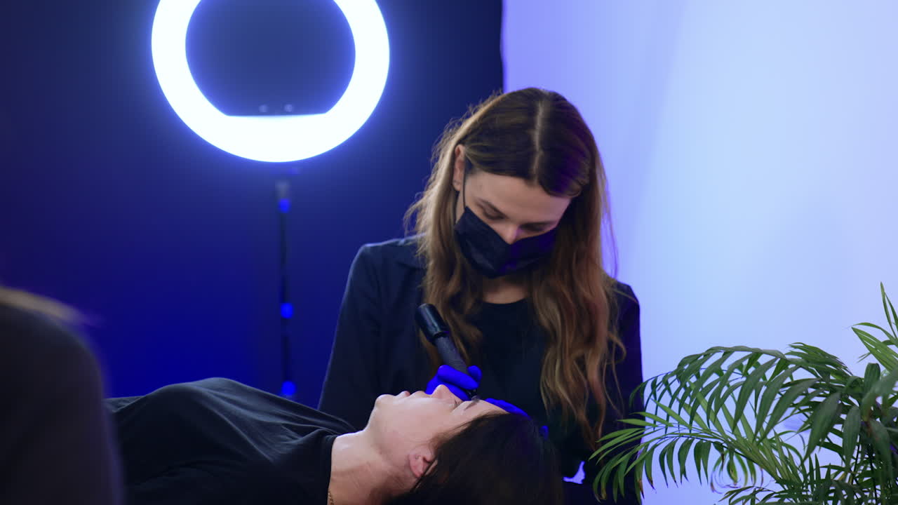Caucasian cosmetologist wearing black mask working in a beauty salon. Specialist performing permanent eyebrow make up.