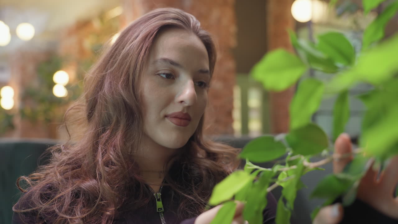 Curly-haired woman in black outfit gently touches vibrant green leaf while standing indoors with warm lighting and blurred background of brick walls, creating a calm natural contrast