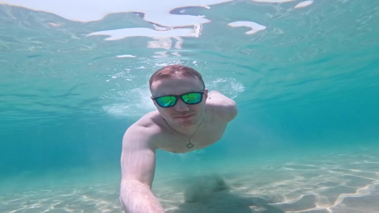 Uomo a torso nudo con occhiali da sole nuota sott'acqua con una action camera nel limpido mare turchese greco, creando un divertente punto di vista (POV) di vacanza