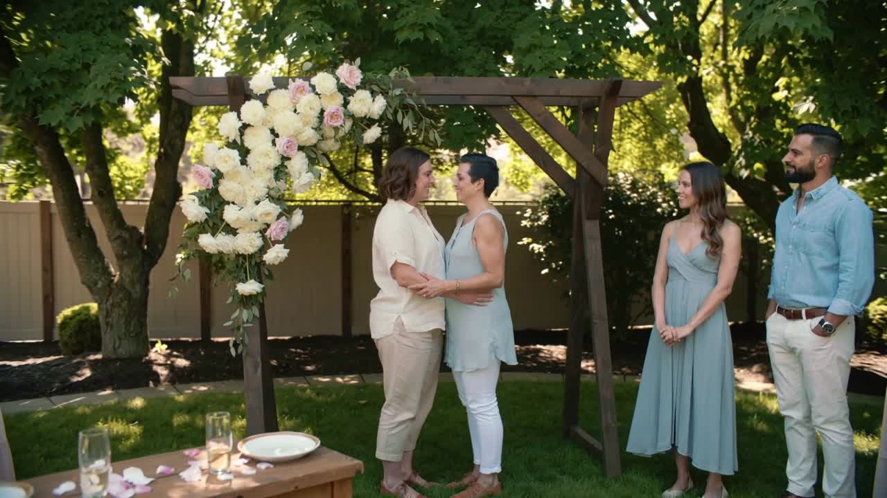 Tenendosi per mano, una coppia in abiti chiari sorride sotto un arco di legno in giardino, con petali, all'inizio della cerimonia.