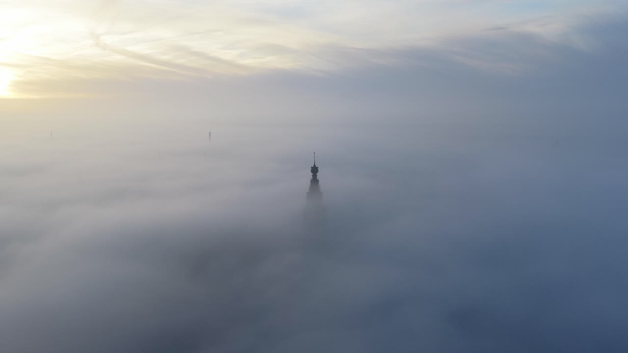 Vista aerea della Stevenskerk che si erge su Nimega durante un'alba nebbiosa, con una leggera foschia che si diffonde tra le stradine e una luce soffusa che colora il cielo. La torre della chiesa emerge dalla foschia.