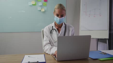 caucasian female doctor wearing mask sitting at desk in meeting room using laptop