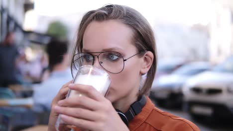 Close-up-footage-of-a-beautiful-blonde-girl-talking-by-mobile-while-drinking-cappuccino-from-glass.-Outdoors,-cafe.-Blurred