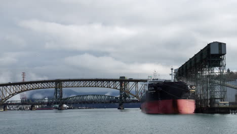 Buque-Portacontenedores-Atracado-En-La-Terminal-De-Viterra-Cascadia-Con-El-Puente-Conmemorativo-De-Los-Trabajadores-Del-Hierro-Y-El-Segundo-Puente-Estrecho-En-El-Fondo-Sobre-La-Entrada-De-Burrard-En-Vancouver,-Columbia-Británica,-Canadá