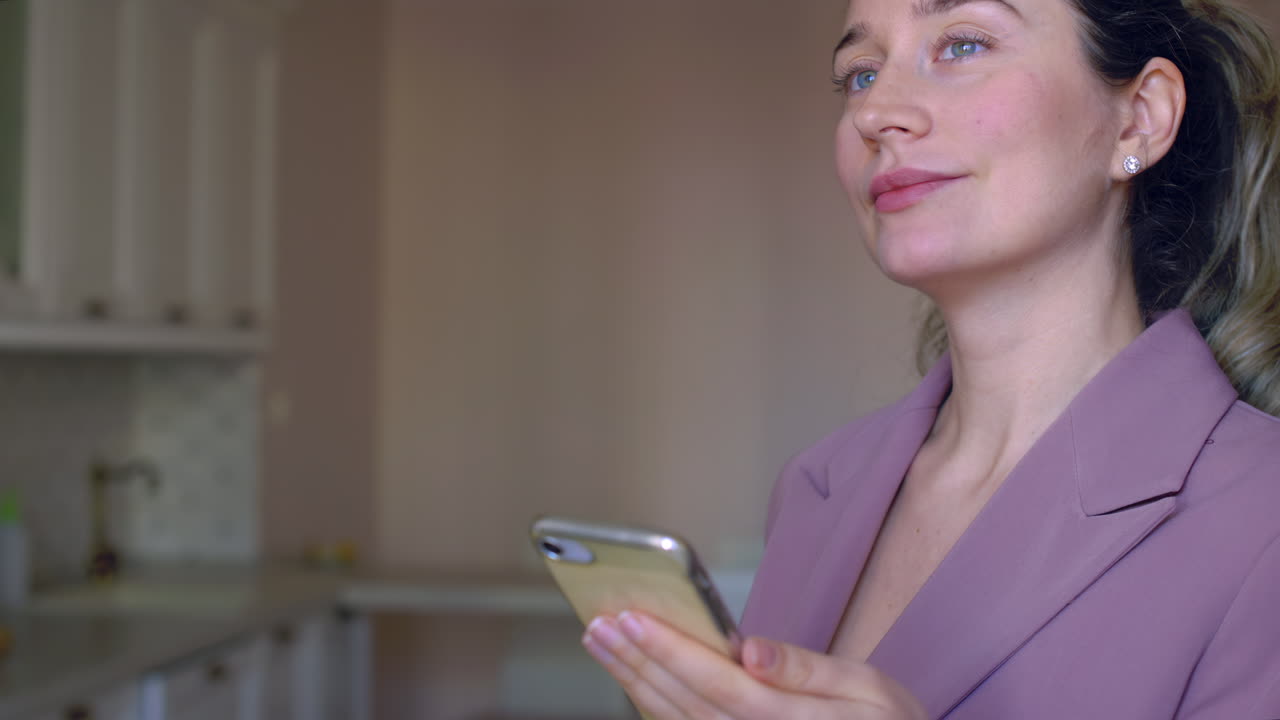 A young woman looks pleased while holding her smartphone in a stylish kitchen. She appears thoughtful and relaxed in her inviting space as sunlight streams in