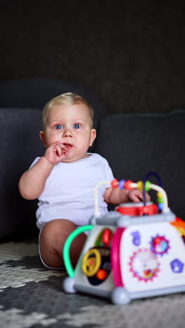 beautiful blond baby sitting on the floor holding a finger in mouth. cute kid looks up not paying attention to a toy in front of him. vertical video.