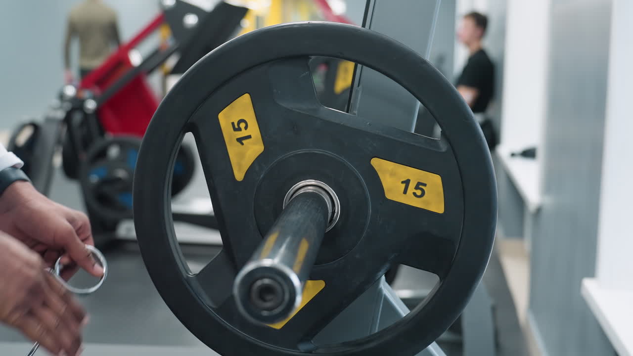 close up of person wearing white sports jacket fitting metal weight plate onto barbell sleeve and securing it with spring clip on rubber gym floor, equipment rack and members visible in background