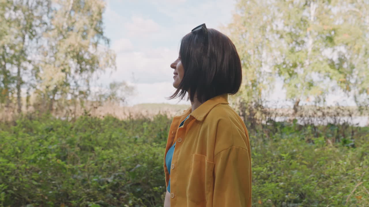 Hvid kvinde smiler under udendørs camping. En munter opdagelsesrejsende i gul skjorte overskuer en åben eng og vender sig mod kameraet. Livlig atmosfære med brise gennem træerne og en lys himmel, venlig og uformel.