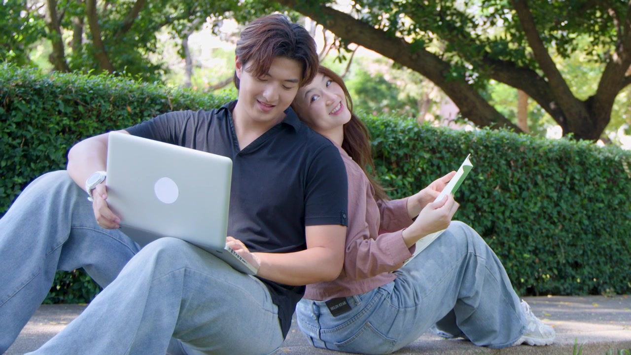 คู่รักหนุ่มสาวนั่งหันหลังชนกัน ใช้แล็ปท็อปและอ่านหนังสือในสวนสาธารณะ