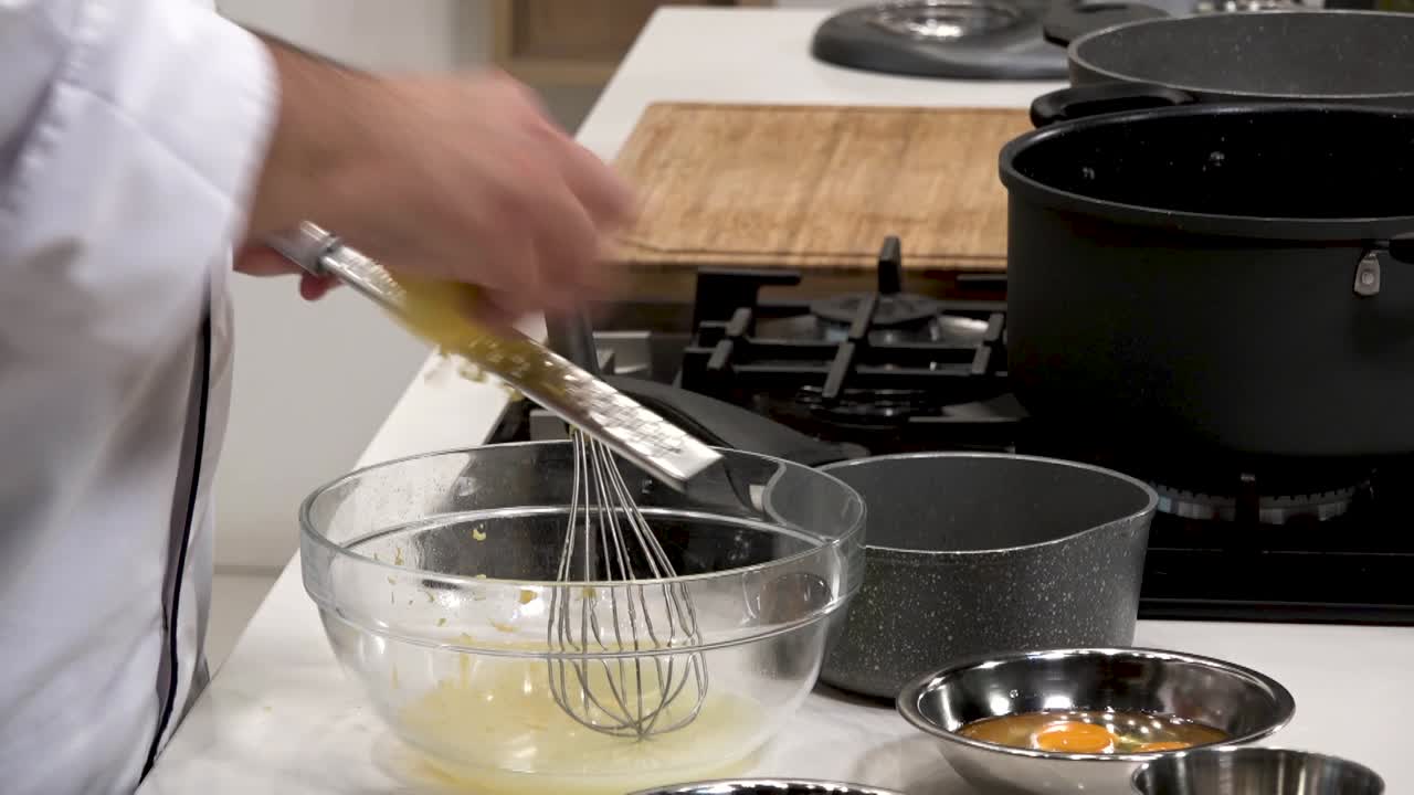 Chef skillfully whisks eggs in a bowl while surrounded by various cooking utensils and pots on a pristine kitchen counter, demonstrating culinary techniques