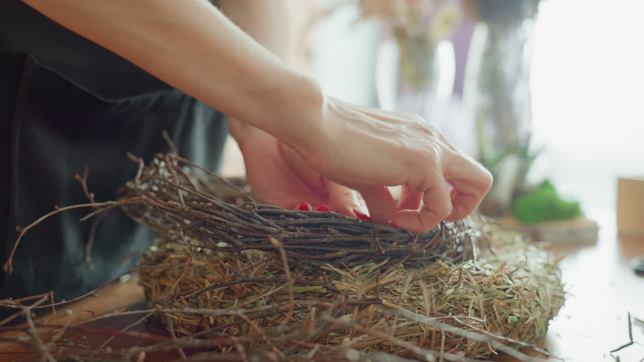 Female florist arranging twig wreath on straw base with delicate hands, showcasing crafting process, rustic natural materials, creative handmade seasonal decoration, and artistic on wooden table indoors