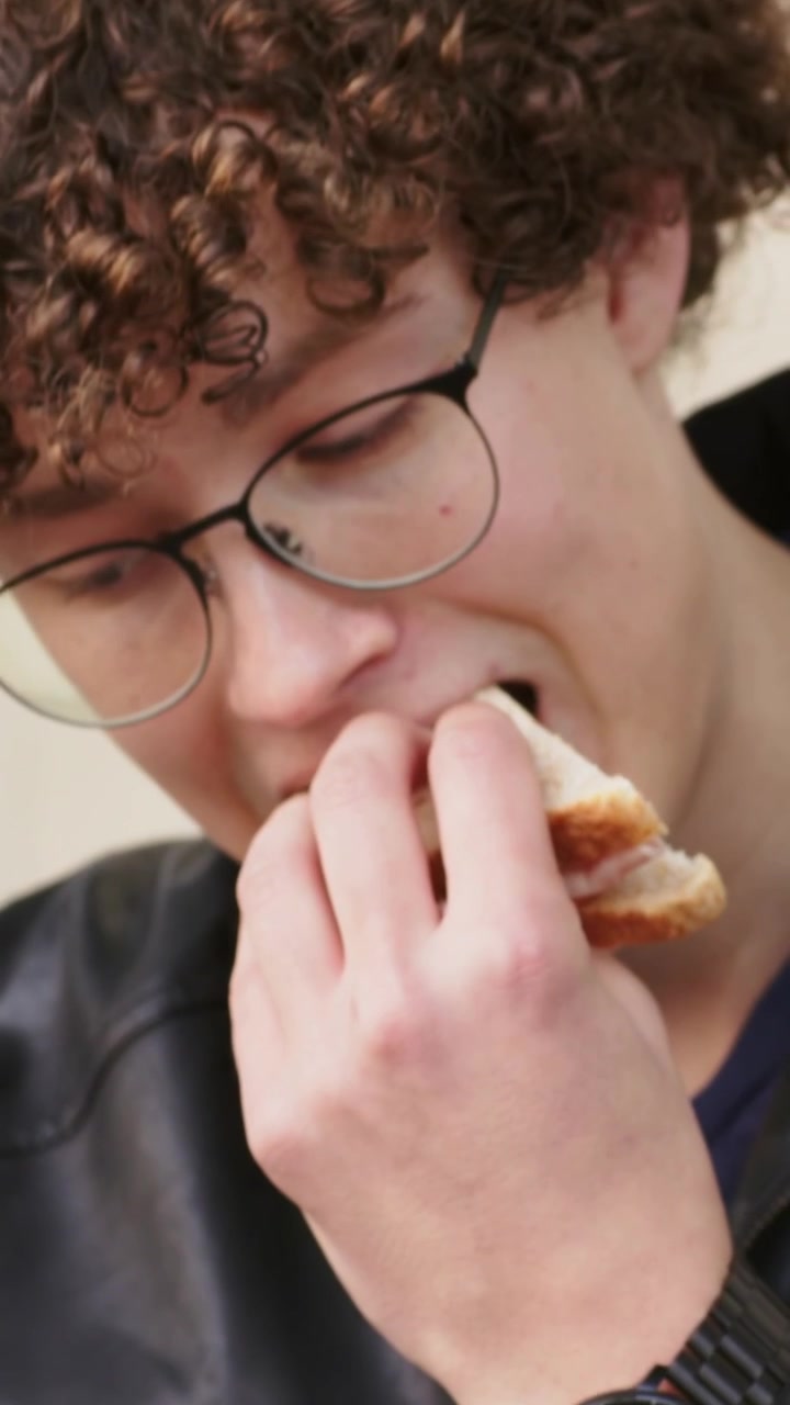 Vertical video: Holding folded sandwich to mouth, man taking bite for snack, with glasses and shirt