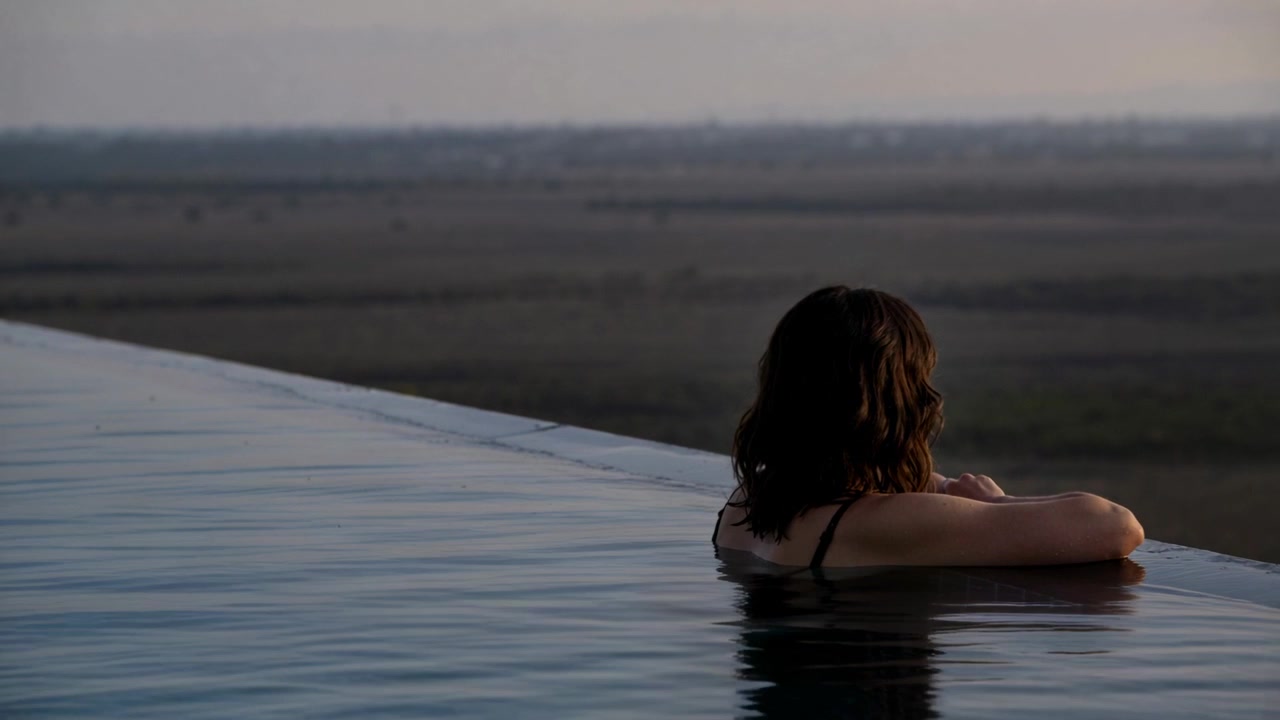 Frau in dunklem Badeanzug beobachtet Sonnenuntergang am fernen Horizont eines Infinity-Pools, mit Freiraum