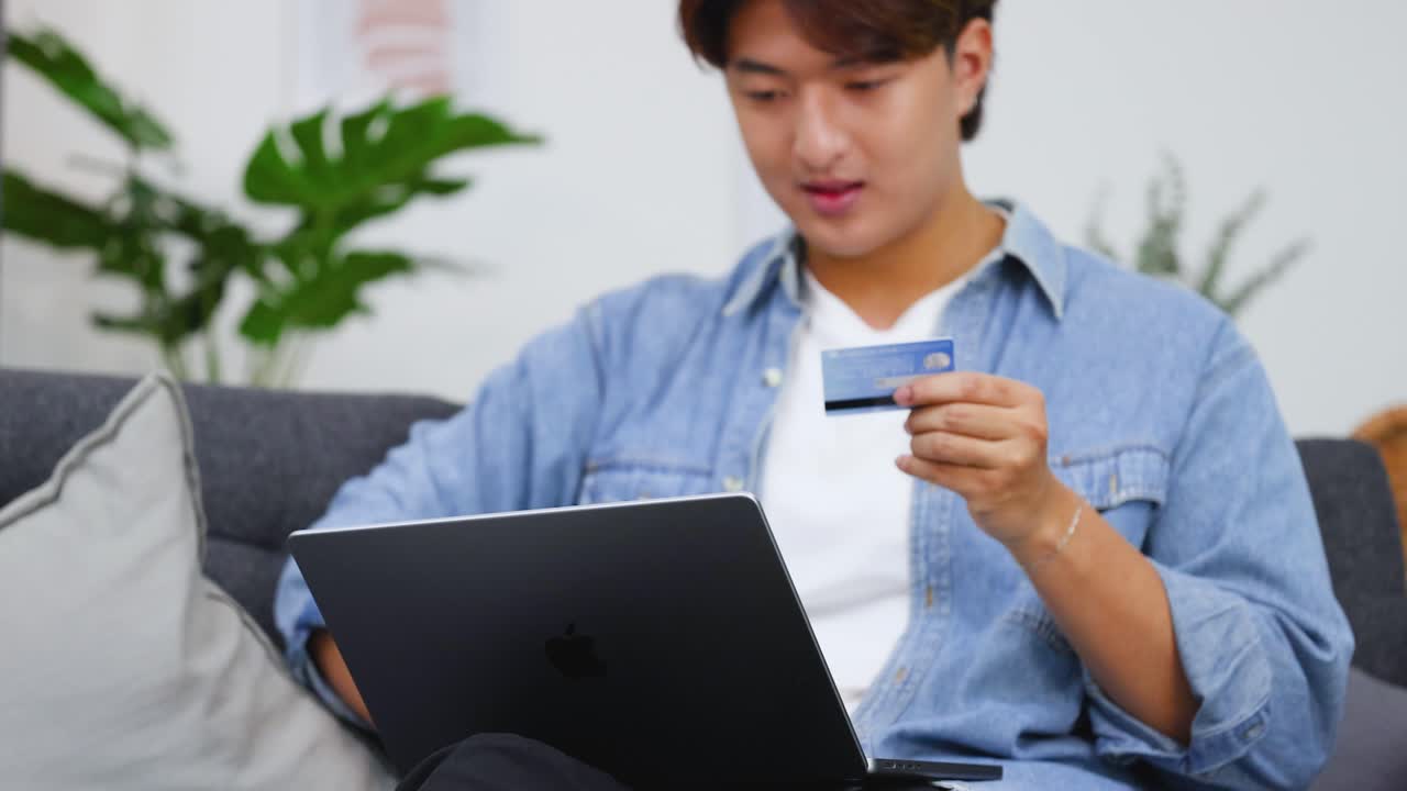 A young man sits on a sofa, entering credit card details into his laptop computer