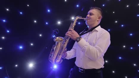tilt up shot of biracial jazz musician in checkered suit and bowtie playing saxophone while sitting against black band member in dimly lit bar, rehearsing and checking sound before live performance