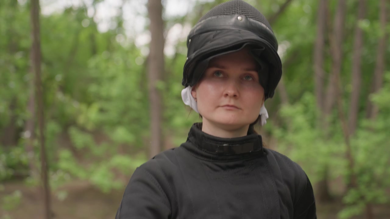 Close up of woman in black fencing gear holding and moving with sword outdoors. subject demonstrates various stances while wearing protective equipment including helmet and padded jacket