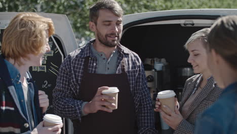 joven barista con barba usando delantal parado frente a su camioneta tomando café con gente joven y charlando