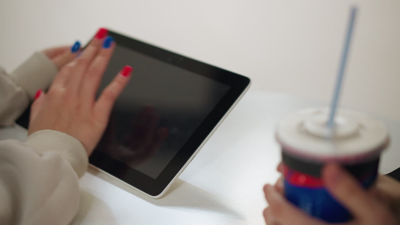 Close up of female hand with red and blue painted nails interacting with tablet while holding drink cup with straw, representing digital lifestyle, online activity and modern technology