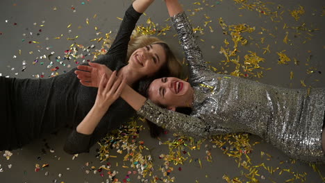 two female friends lying on the floor full of confetti after a glamorous new years eve party, laughing and dancing hand in hand