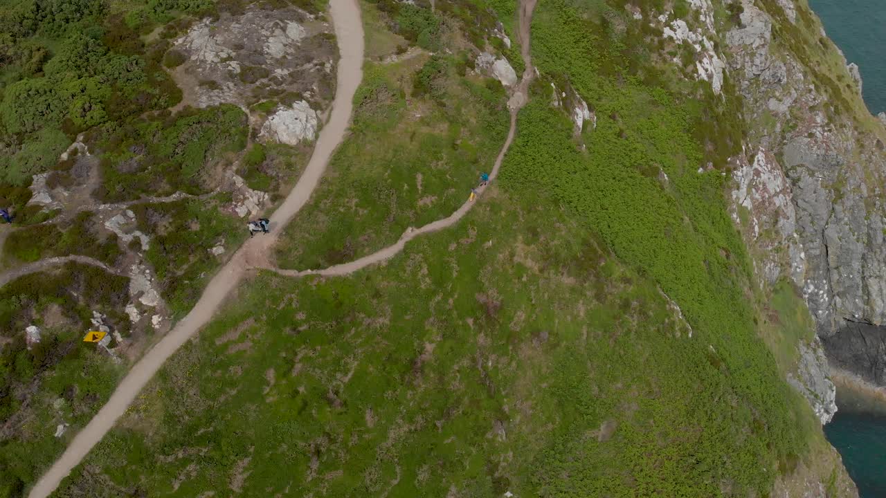imagen de arriba hacia abajo de turistas caminando por una montaña verde junto al océano en un día soleado