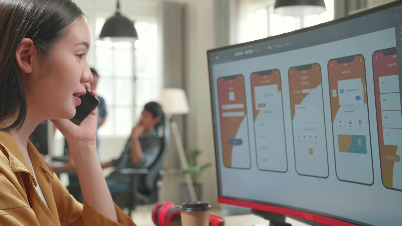 Asian Female Mobile Application Developer Works And Talking On Mobile Phone With Graphics On Her Personal Computer In Office