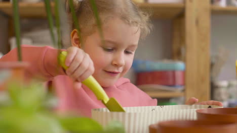 close up view of little blonde girl preparing the soil in a pot sitting at a table where is plants in a craft workshop