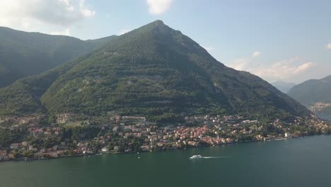 a large green mountain next to a lake with a big boat driving by in italy, como lake