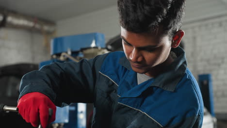 mechanic in blue uniform and red gloves tightening bolts on car engine in workshop, background features industrial tools, open car hood, and mechanical equipment