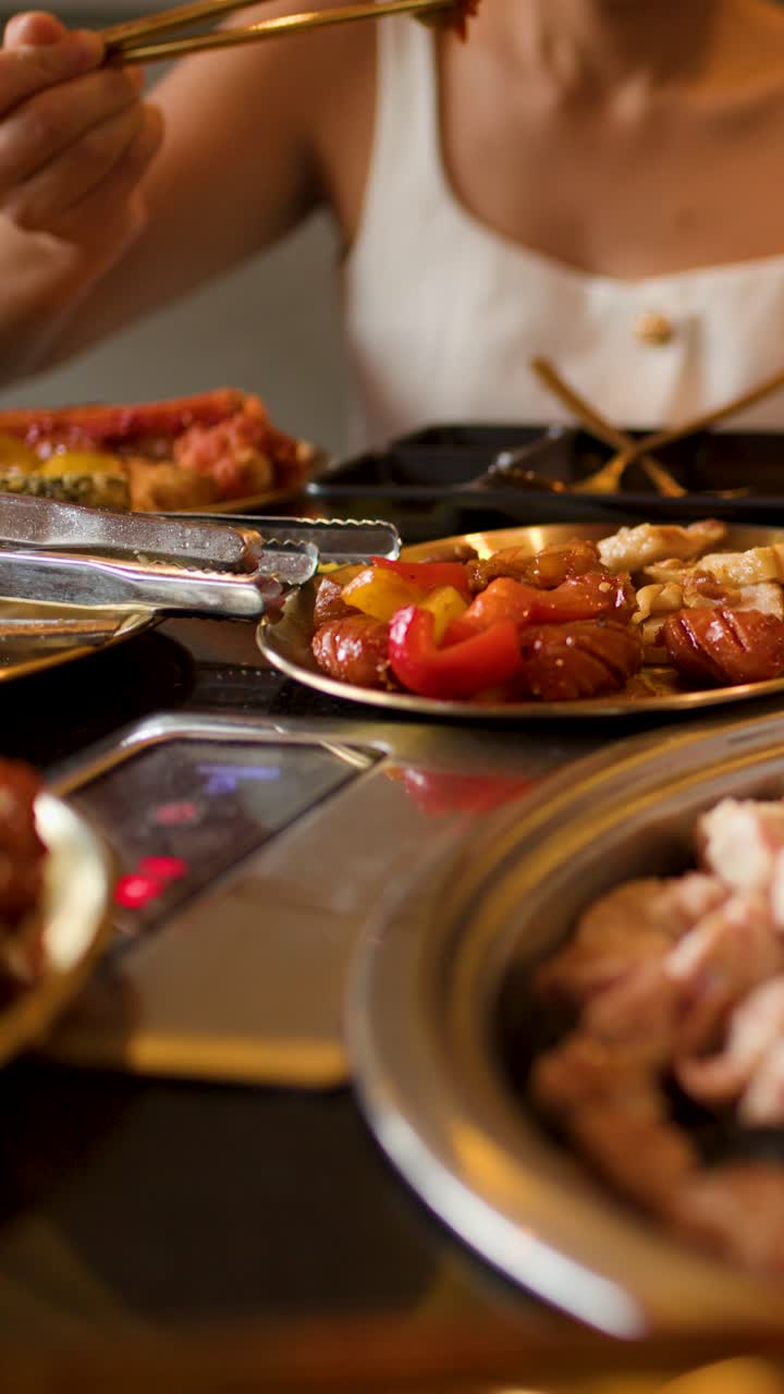Woman uses chopsticks to cook and eat assorted meats at tabletop grill, warm ambient lighting